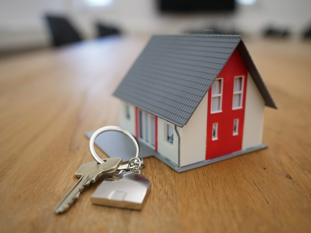 A small model house with a red vertical stripe sits next to a house key on a wood table.